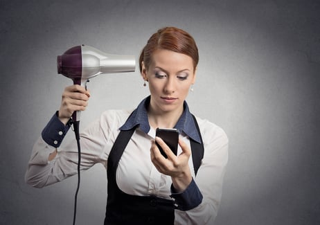 Closeup portrait business woman deal maker reading news on smartphone looking at mobile phone holding hairdryer isolated grey wall background. Human face expression. Busy life of corporate executive