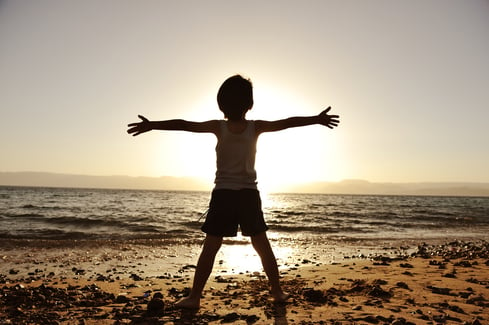 Silhouette of child on the beach, holding his hands up, hugging the sun