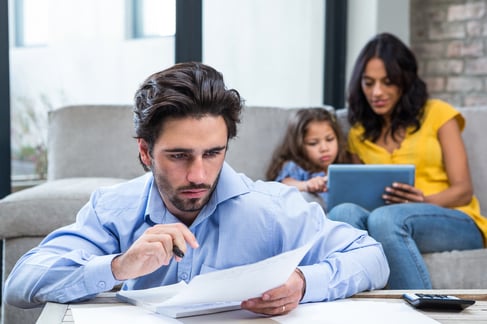 Thoughtful father paying bills in living room while wife and daughter are on the sofa